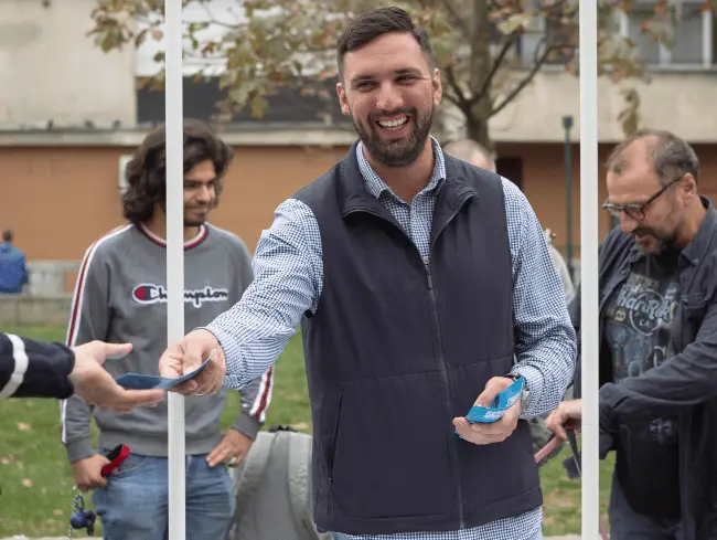 Muškarac deli letke u parku, nasmejan, okružen ljudima.