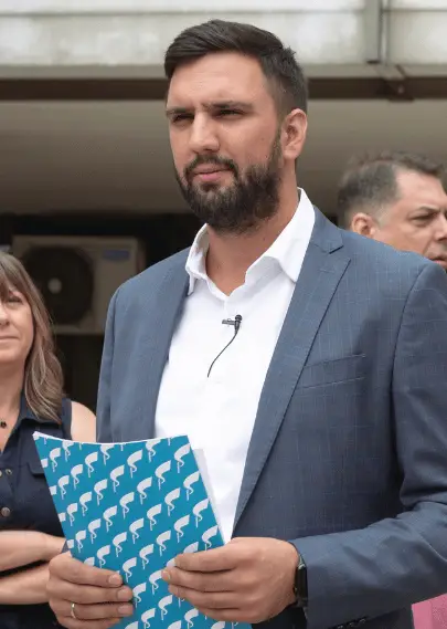 Muškarac drži plavu fasciklu i govori na konferenciji.